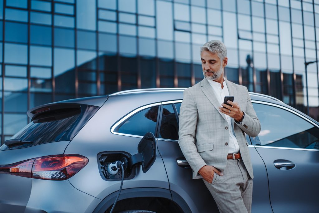 Businessman holding smartphone while charging car at electric vehicle charging station, closeup.